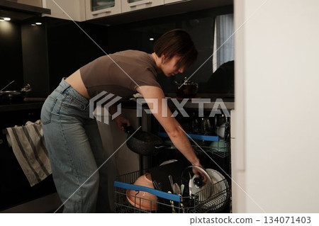 A woman loads dishes and a pot into a dishwasher, completing a simple household chore in a modern kitchen 134071403