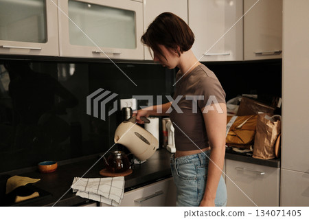 A woman pours hot water from an electric kettle into a glass teapot, preparing a warm drink in a calm kitchen setting A woman pours hot water from an electric kettle into a glass teapot, preparing a warm drink in a calm kitchen setting 134071405