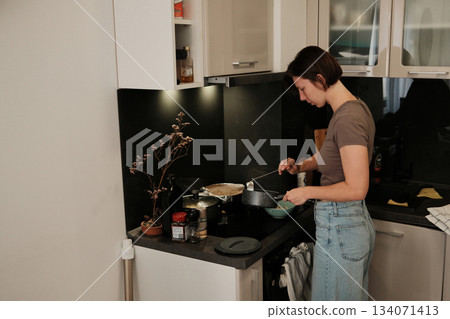 A woman prepares food in a modern kitchen, carefully ladling hot soup into a bowl as part of her daily cooking routine 134071413