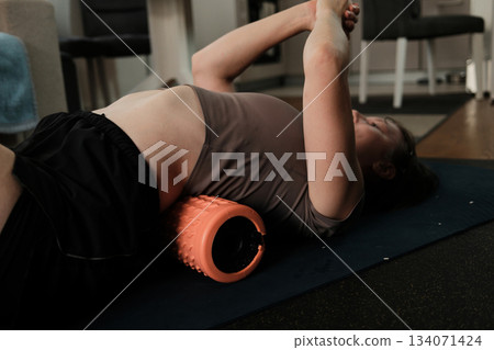 A woman lies on a textured foam roller during a home recovery session, gently loosening back muscles and relieving tension 134071424
