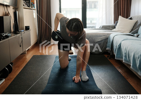 A woman performs a kneeling dumbbell row on a mat during a home workout, focusing on strength, form, and controlled movement in a calm indoor setting A woman performs a kneeling dumbbell row on a mat during a home workout, focusing on strength, form, and controlled movement in a calm indoor setting 134071428