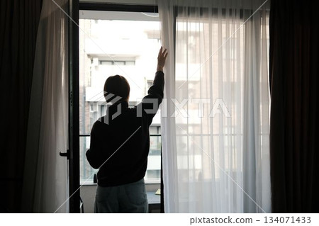 A woman stands near an open balcony door, gently pulling back sheer curtains to let fresh daylight illuminate the room A woman stands near an open balcony door, gently pulling back sheer curtains to let fresh daylight illuminate the room 134071433