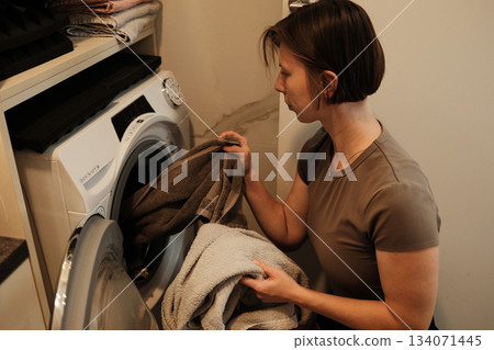 A woman places towels into a washing machine, continuing her household chores in a warm and organized home setting A woman places towels into a washing machine, continuing her household chores in a warm and organized home setting 134071445