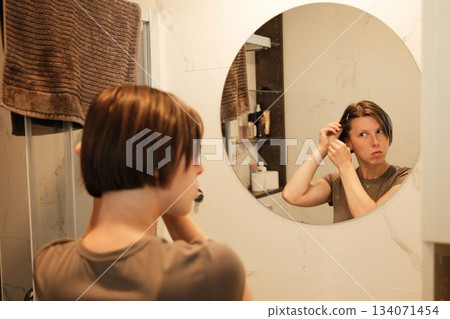 A woman uses a hairpin while looking into a mirror, adjusting her hairstyle as part of her organized morning grooming routine A woman uses a hairpin while looking into a mirror, adjusting her hairstyle as part of her organized morning grooming routine 134071454