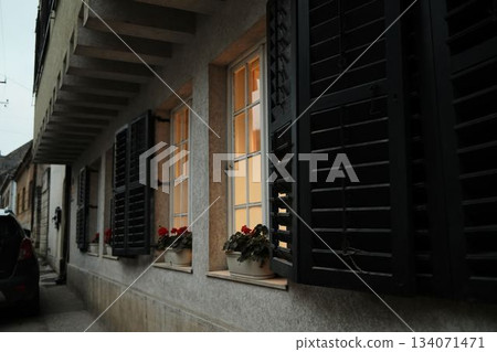 Warm light shines through open shutters and decorated windows on a quiet street in Sremski Karlovci, Serbia. Flower pots add a homely and inviting feeling Warm light shines through open shutters and decorated windows on a quiet street in Sremski Karlovci, Serbia. Flower pots add a homely and inviting feeling 134071471