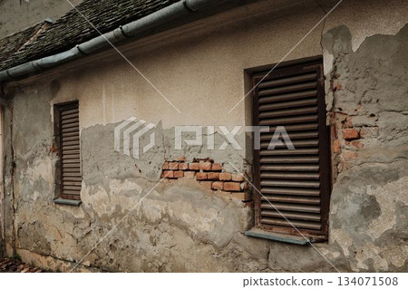 A weathered house wall shows cracked plaster and exposed bricks beneath wooden shutters. The image reflects decay and rural simplicity 134071508