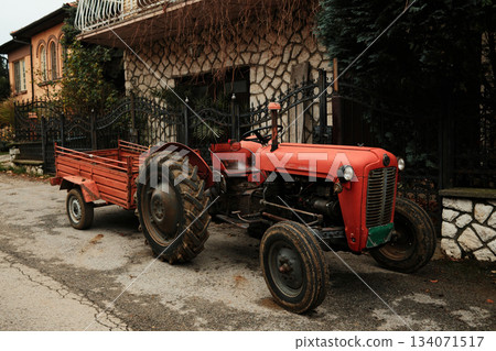 A red tractor with a small trailer stands on a quiet village road. The scene reflects rural work and agricultural lifestyle A red tractor with a small trailer stands on a quiet village road. The scene reflects rural work and agricultural lifestyle 134071517