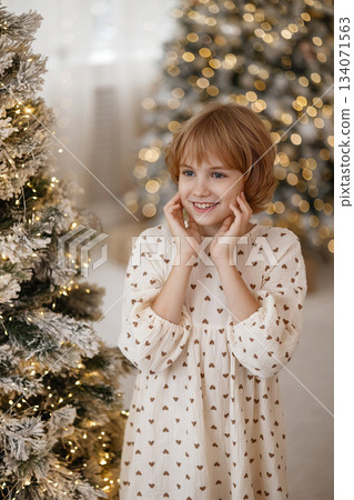 Caucasian blonde girl smiling and holding face near Christmas tree in cozy home interior Caucasian blonde girl smiling and holding face near Christmas tree in cozy home interior 134071563
