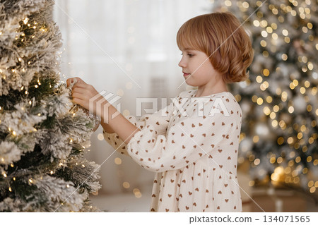 Side view of Caucasian blonde girl decorating Christmas tree in cozy room Side view of Caucasian blonde girl decorating Christmas tree in cozy room 134071565