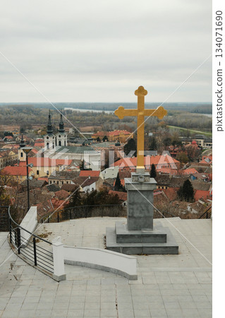 Wide view of Sremski Karlovci with golden cross in the foreground, red rooftops, churches, and the Danube in the distance. A cultural landscape under soft clouds 134071690
