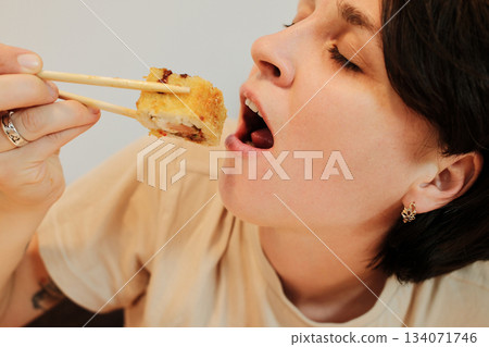 A close-up view of a woman opening her mouth to eat a fried sushi roll held with chopsticks. The shot focuses on appetite, taste, and an expressive dining moment 134071746