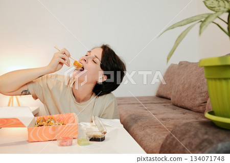 A woman leans toward the table and takes a large bite of fried sushi with chopsticks. The scene emphasizes appetite, strong flavor, and casual dining at home A woman leans toward the table and takes a large bite of fried sushi with chopsticks. The scene emphasizes appetite, strong flavor, and casual dining at home 134071748