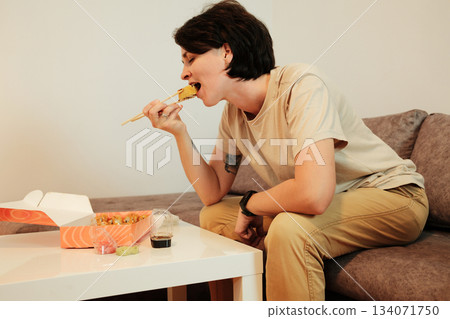 A woman sits on a sofa and eats fried sushi with chopsticks from a delivery box. The scene highlights casual dining, comfort, and modern home lifestyle with Asian food A woman sits on a sofa and eats fried sushi with chopsticks from a delivery box. The scene highlights casual dining, comfort, and modern home lifestyle with Asian food 134071750
