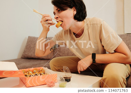 A woman sits on a sofa and eats fried sushi with chopsticks from a delivery box. The scene highlights casual dining, comfort, and modern home lifestyle with Asian food A woman sits on a sofa and eats fried sushi with chopsticks from a delivery box. The scene highlights casual dining, comfort, and modern home lifestyle with Asian food 134071763
