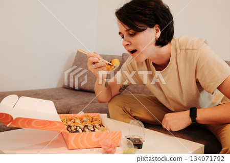 A woman sits on a sofa and eats fried sushi with chopsticks from a delivery box. The scene highlights casual dining, comfort, and modern home lifestyle with Asian food A woman sits on a sofa and eats fried sushi with chopsticks from a delivery box. The scene highlights casual dining, comfort, and modern home lifestyle with Asian food 134071792