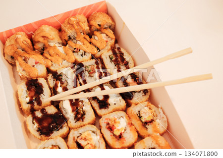 Chopsticks rest on a box of assorted fried sushi rolls topped with sauces and crispy textures. The warm scene highlights Asian flavors and convenient modern dining Chopsticks rest on a box of assorted fried sushi rolls topped with sauces and crispy textures. The warm scene highlights Asian flavors and convenient modern dining 134071800