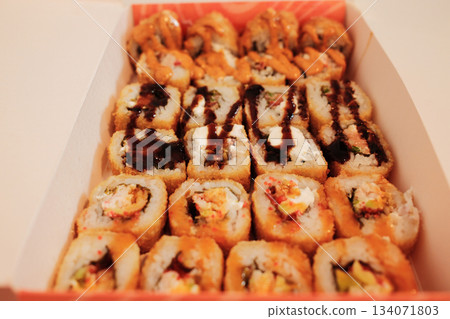 A close view of assorted fried sushi rolls arranged in a delivery box, topped with sauces and crispy textures. The image highlights variety, richness, and modern takeaway cuisine A close view of assorted fried sushi rolls arranged in a delivery box, topped with sauces and crispy textures. The image highlights variety, richness, and modern takeaway cuisine 134071803