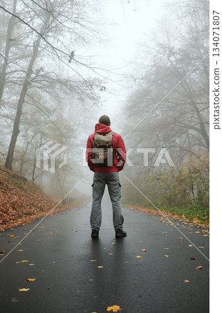 Man with backpack stands still on wet autumn road surrounded by fog and fallen leaves. Back rear view. Quiet and cinematic scene Man with backpack stands still on wet autumn road surrounded by fog and fallen leaves. Back rear view. Quiet and cinematic scene 134071807