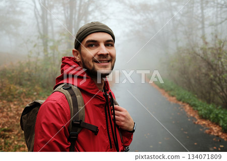 Happy male traveler in red jacket smiling on quiet misty forest road during autumn walk. Solo travel concept Happy male traveler in red jacket smiling on quiet misty forest road during autumn walk. Solo travel concept 134071809