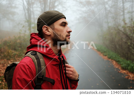 Side view of traveler with backpack gazing into foggy autumn forest road. Calm and thoughtful atmosphere 134071810