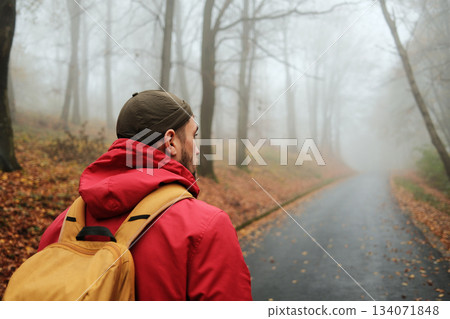 Back rear view of male traveler in red jacket and backpack looking at foggy autumn forest road 134071848