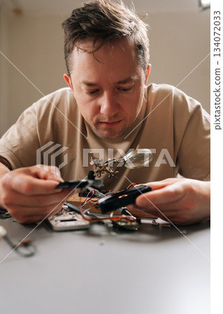 Vertical portrait of serious focused technician examining complex circuit board with magnifying glass, performing detailed electronic repair work, problem-solving, and developing new technology. Vertical portrait of serious focused technician examining complex circuit board with magnifying glass, performing detailed electronic repair work, problem-solving, and developing new technology. 134072033