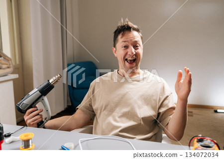 Portrait of happy man engaging in do-it-yourself project, showcasing excitement and enthusiasm holding heat gun at a workbench, illustrating passion for home repairs and electronic work. 134072034