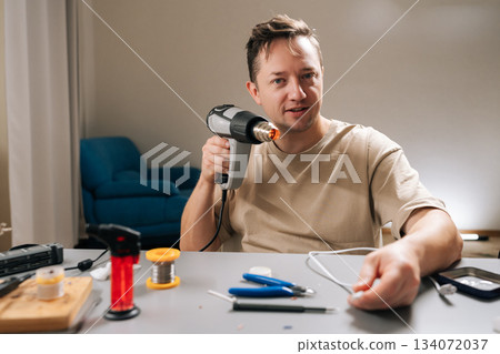 Professional repairman working on damaged electronic cable, applying heat with heat gun to shrink tubing or solder connecting wires, signifying diligent repair work and technical expertise. Professional repairman working on damaged electronic cable, applying heat with heat gun to shrink tubing or solder connecting wires, signifying diligent repair work and technical expertise. 134072037