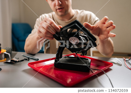 Man working with computer parts, holding cooling system fan over radiator, focusing on te intricate process of electronics assembly and repair. Concept of hardware, repairing, upgrade and technology. Man working with computer parts, holding cooling system fan over radiator, focusing on te intricate process of electronics assembly and repair. Concept of hardware, repairing, upgrade and technology. 134072039