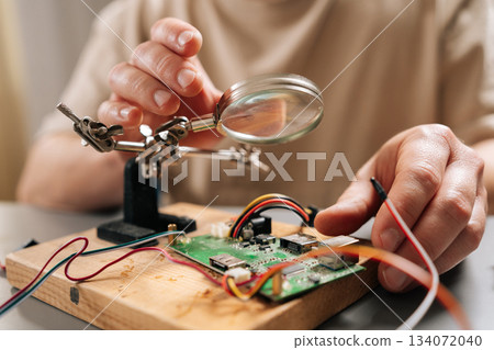 Close-up hands of technician focusing on intricate details of circuit board on wooden base with magnifying glass, using helping hands tools and wires during electronic repair or assembly process. Close-up hands of technician focusing on intricate details of circuit board on wooden base with magnifying glass, using helping hands tools and wires during electronic repair or assembly process. 134072040