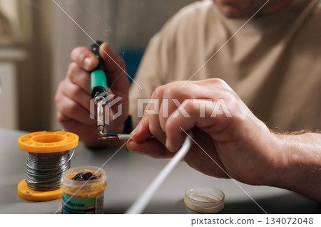 Close-up cropped shot of person soldering wires using electric soldering iron, creating smoke, repairing damaged electrical cable, performing electronic circuit work in modern workshop environment. Close-up cropped shot of person soldering wires using electric soldering iron, creating smoke, repairing damaged electrical cable, performing electronic circuit work in modern workshop environment. 134072048