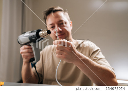 Focused repairman working on damaged electronic cable, applying heat with heat gun to shrink tubing or solder connecting wires, signifying diligent repair work and technical expertise. Focused repairman working on damaged electronic cable, applying heat with heat gun to shrink tubing or solder connecting wires, signifying diligent repair work and technical expertise. 134072054