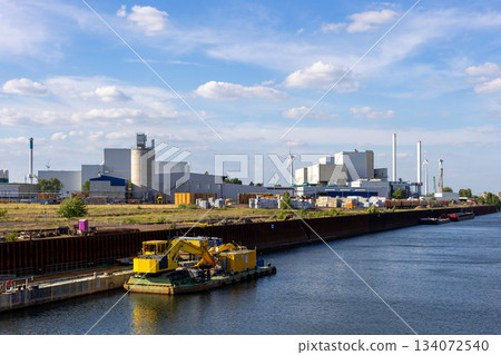 Industrial harbor area in Magdeburg shows factories, storage yards and wind turbines near calm water. Bright machinery and barges highlight active logistics under a clear blue sky Industrial harbor area in Magdeburg shows factories, storage yards and wind turbines near calm water. Bright machinery and barges highlight active logistics under a clear blue sky 134072540