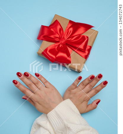 A woman's hands crossed in a gesture of refusal over a gift box wrapped in craft paper with a red bow. Against a blue background. A woman's hands crossed in a gesture of refusal over a gift box wrapped in craft paper with a red bow. Against a blue background. 134072743