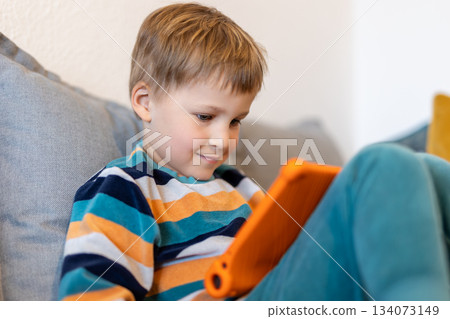 Portrait of cute adorable blond little kid boy enjoy sitting on home sofa surf web play game or watching TV show movies using tablet. Children smart technology usage concept. Child gadget addiction Portrait of cute adorable blond little kid boy enjoy sitting on home sofa surf web play game or watching TV show movies using tablet. Children smart technology usage concept. Child gadget addiction 134073149