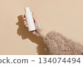 A womans hand in a sweater holds a bottle of cream on a beige background. Body care beauty treatment 134074494