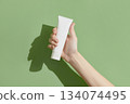 A womans hand holding a white cosmetic tube on a green background 134074495