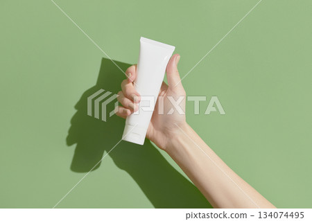 A womans hand holding a white cosmetic tube on a green background A womans hand holding a white cosmetic tube on a green background 134074495