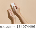 A womans hands holding a white cosmetic tube on a beige background. Beauty treatment concept 134074496
