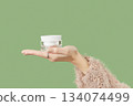 A womans hand in a sweater holds a jar of cream on a green background. Self care beauty treatment 134074499