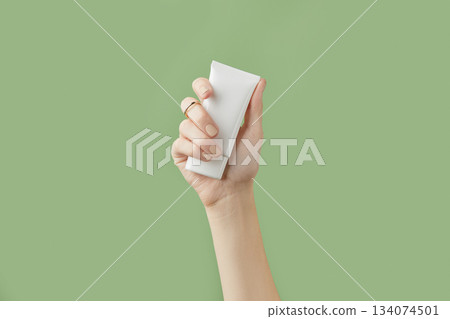 A womans hand holding a white cosmetic tube on a green background A womans hand holding a white cosmetic tube on a green background 134074501