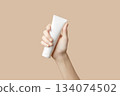 A womans hand holding a white cosmetic tube on a beige background. Care products 134074502