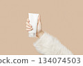 A womans hand in a sweater holds a tube of cream on a beige background. Body care beauty treatment 134074503