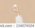 A womans hand in a sweater holds a jar of cream on a beige background. Body care beauty treatment 134074504