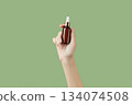 A womans hand holding a brown bottle of serum on a green background 134074508