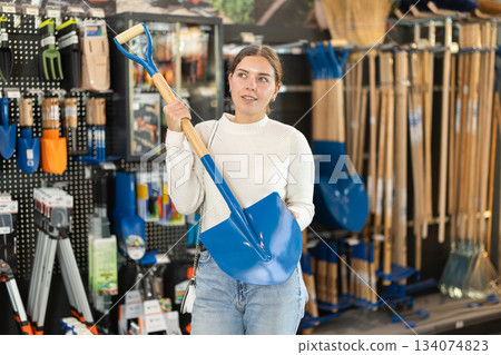Girl in hardware store examines shovel and spade. Girl in hardware store examines shovel and spade. 134074823