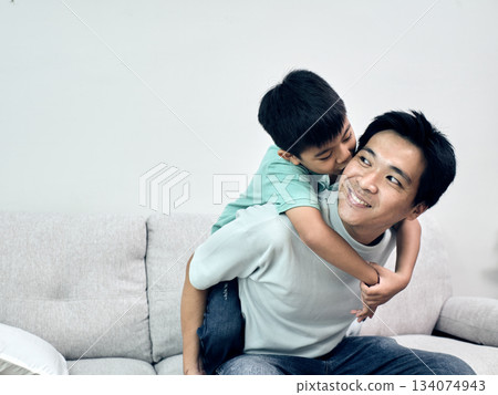 Asian father smiles as his son hugs and kisses him from behind on the couch in a joyful home moment Asian father smiles as his son hugs and kisses him from behind on the couch in a joyful home moment 134074943