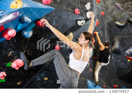 Sporty strong young female exercising in boulder climbing hall without rope, reaching new results, enjoying new challenges in amusement park Sporty strong young female exercising in boulder climbing hall without rope, reaching new results, enjoying new challenges in amusement park 134074957