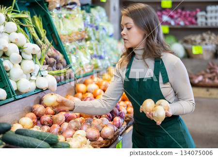 Young female seller putting onions in vegetable basket in grocery market 134075006