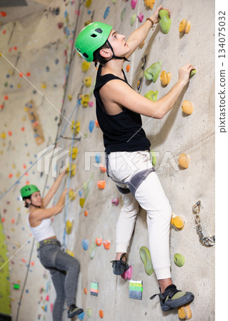 young positive man, insured by trainer, moving up artificial climbing wall indoors 134075032